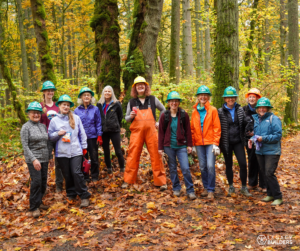 A group of women volunteers stand together smiling on a leaf covered trail.