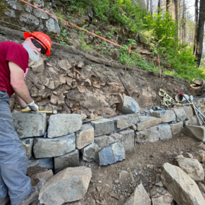 A volunteer looks back at the camera while constructing a large rock wall.