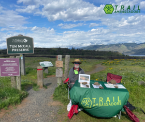 A Trail Ambassador sits behind a table of resources in front of a trailhead sign for Tom McCall.